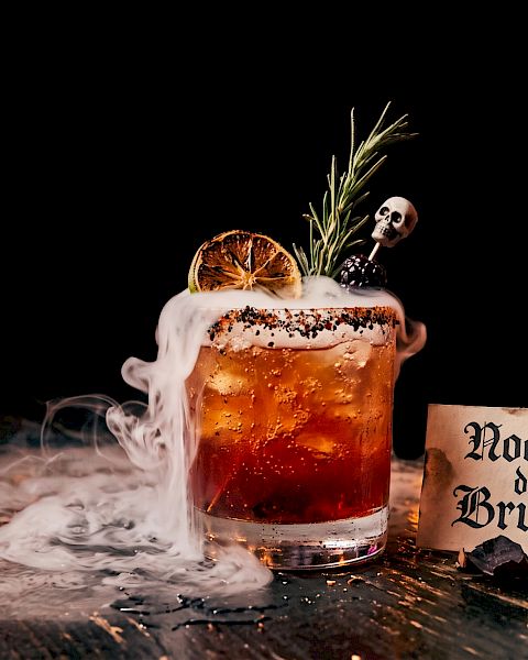 A Halloween &ldquo;night of witches&rdquo; beer: a foamy glass with smoke, orange slice, rosemary, skull garnish, and a &ldquo;Noche de Brujas&rdquo; sign.