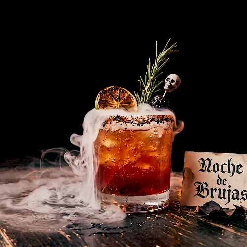 A Halloween “night of witches” beer: a foamy glass with smoke, orange slice, rosemary, skull garnish, and a “Noche de Brujas” sign.