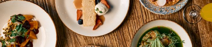 A spread of elegant small plates: a sushi-tinish roll, a green pesto pasta, a taco-like dish, a colorful salad, and a dessert bowl, all on a wooden table, with drinks nearby.